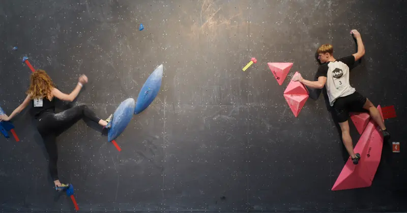 Twee mensen zijn aan het boulderen op een indoor-klimwand, elk op grepen van een andere kleur; de ene zit op blauwe grepen aan de linkerkant, de andere op roze grepen aan de rechterkant.